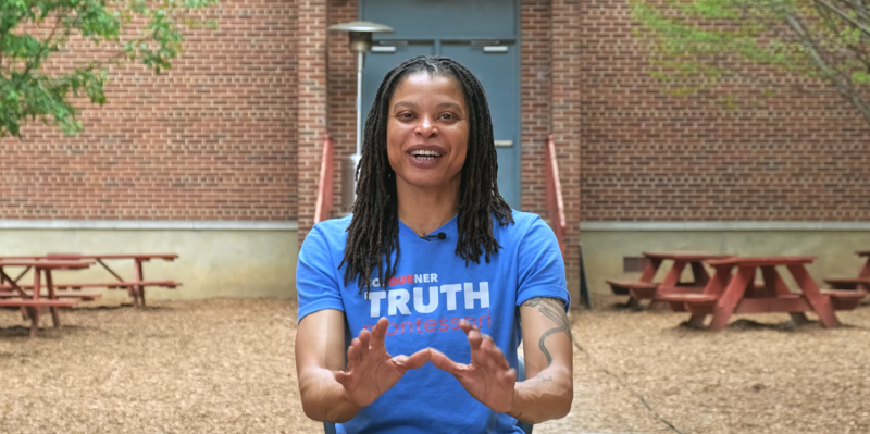 A woman with dreadlocks and a blue t-shirt stands in front of a brick building. She is gesturing with her hands, and there are picnic tables visible in the background. The area appears to be an outdoor courtyard or patio. The woman's t-shirt has some text on it, but it's not entirely clear what it says.